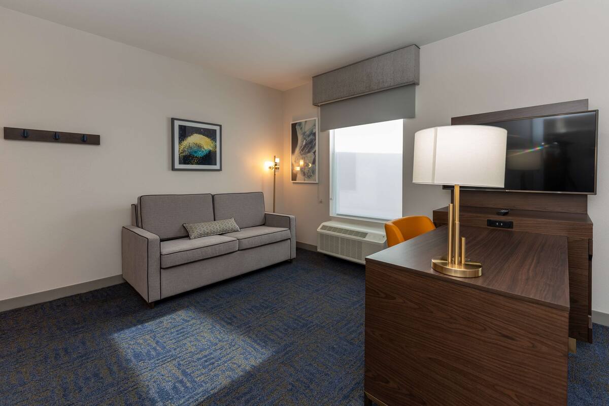 A hotel room with a grey sofa, a wooden desk, a flat-screen TV, and a window with blinds.