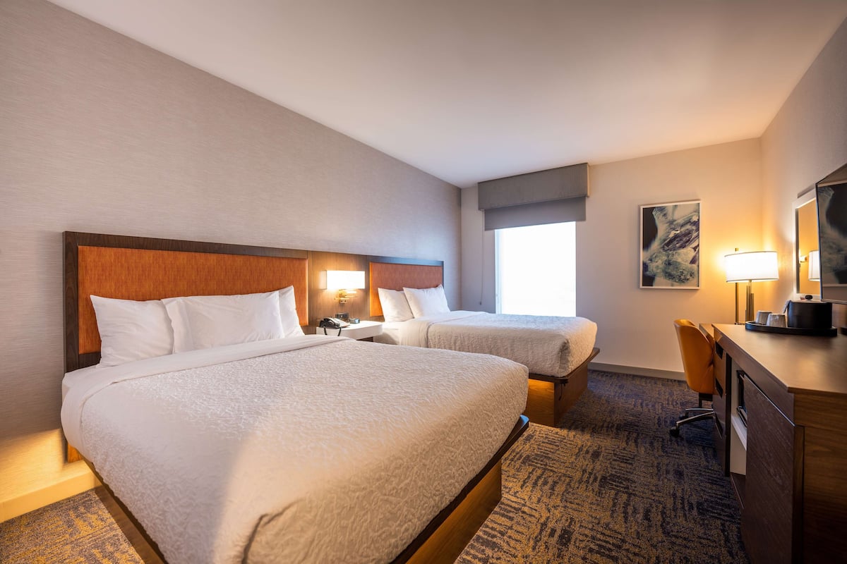 A hotel room with two beds, a desk, a chair, a lamp, and a painting on the wall.