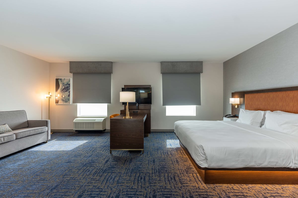 A hotel room with a large bed, a desk with a lamp, a sofa, and a view of the city.
