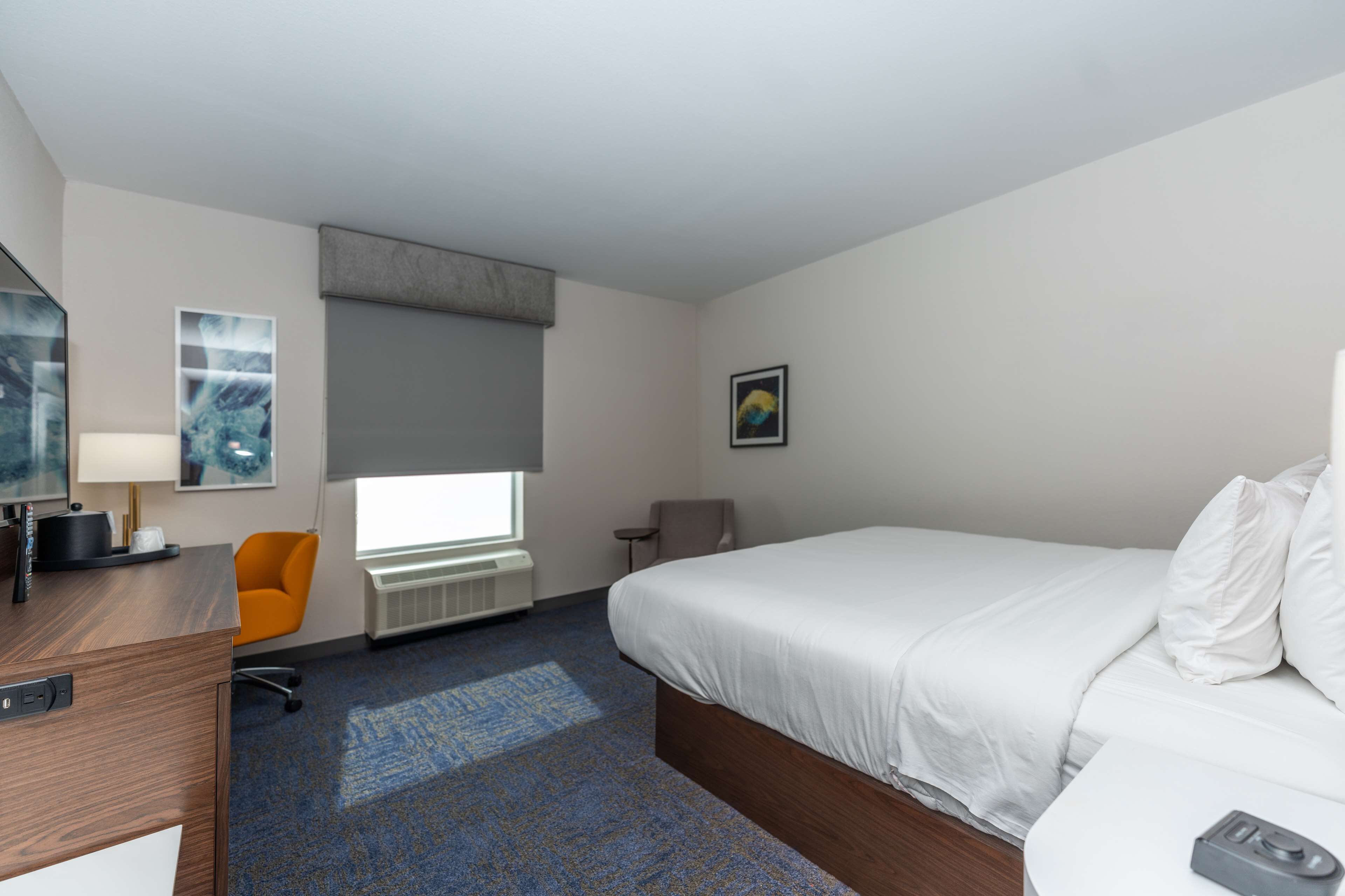 A hotel room with a bed, a desk, a chair, a window, and artwork on the walls.