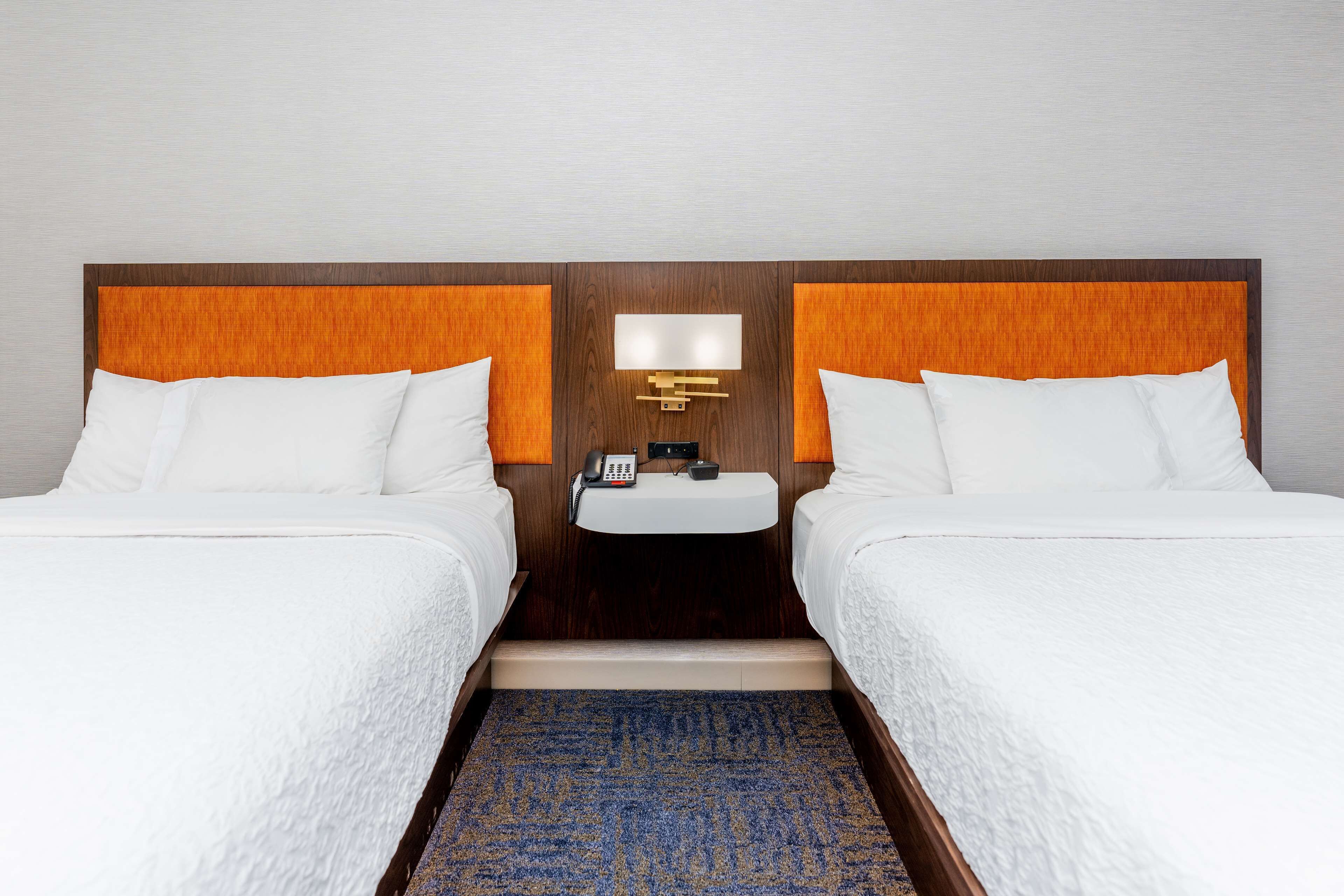 A hotel room with two beds, a nightstand with a lamp, and a blue patterned rug.