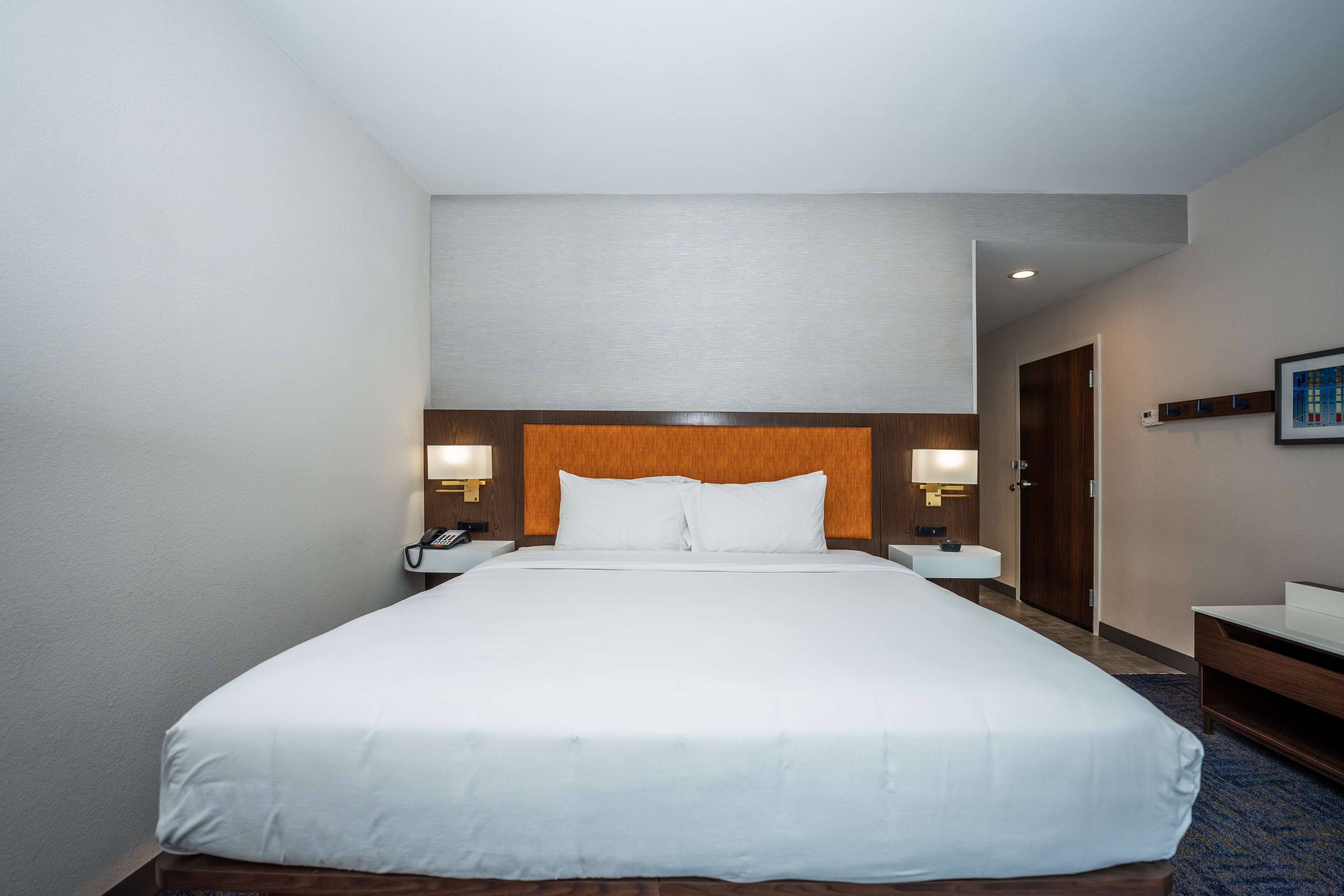 Habitación de hotel con una cama grande, mesitas de noche con lámparas, cabecera de madera, un pequeño escritorio y un cuadro en la pared.