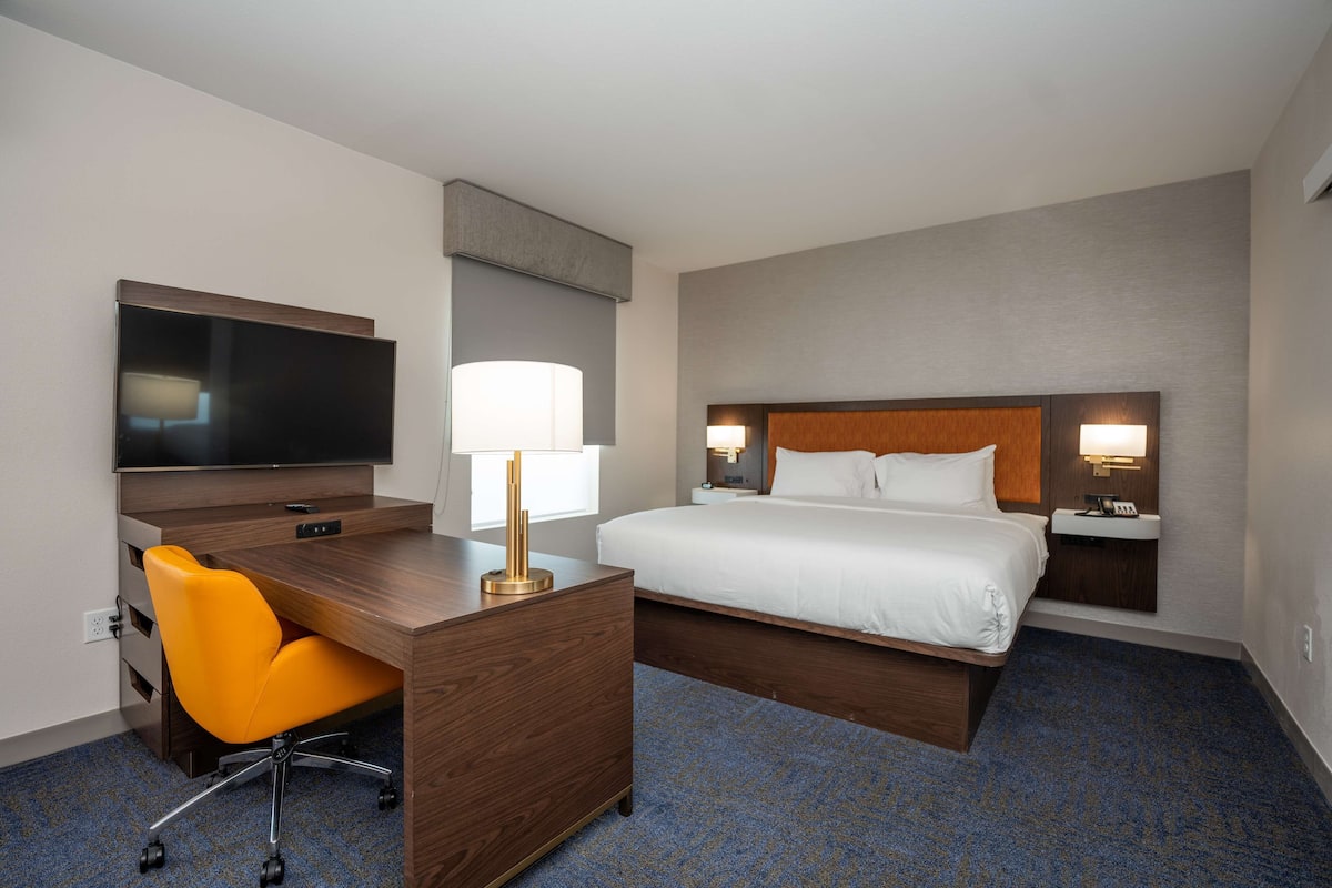 A hotel room with a wooden desk, an orange chair, a bed with white linens, and a flat-screen TV mounted on the wall.