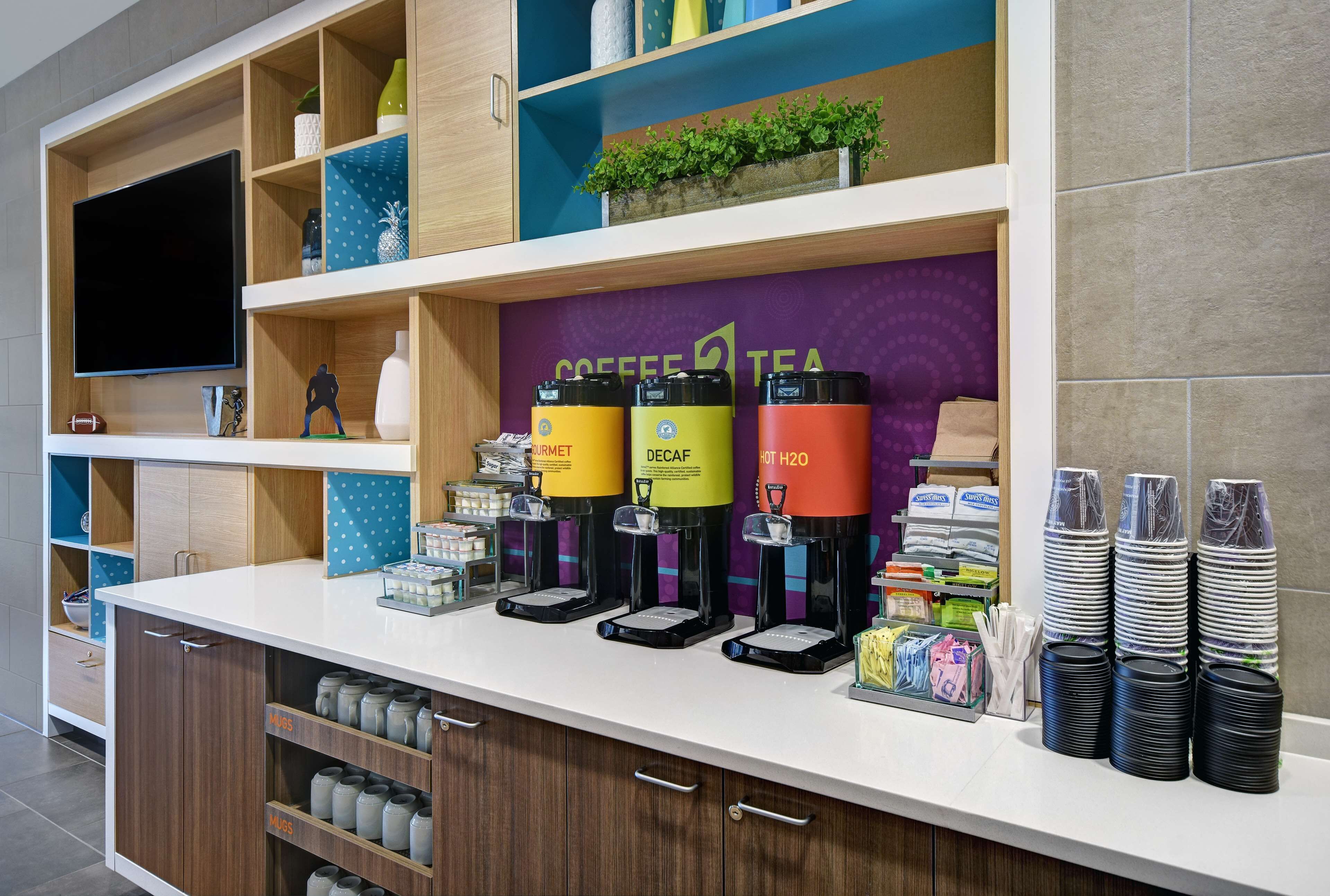 Un espace dédié au café et au thé, équipé de trois distributeurs étiquetés « CAFÉ », « CAFÉ DÉCAFÉINÉ » et « EAU CHAUDE », ainsi que de piles de tasses noires.