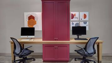 A workspace with two desks, each with a computer monitor, keyboard, and mouse. There are two office chairs, a red cabinet, and artwork on the wall.