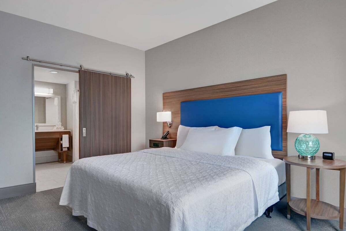 A hotel room with a large bed, a wooden headboard, two bedside tables with lamps, a bathroom visible through a sliding door, and a small round table with a lamp.
