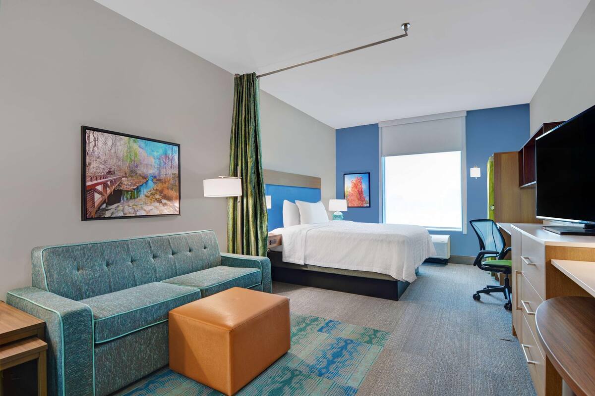 A hotel room with a sofa, bed, desk, and a painting on the wall.