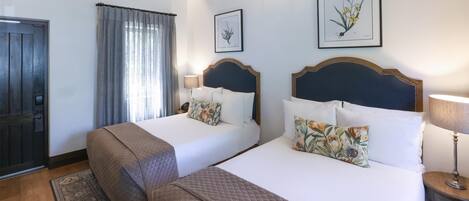 Habitación de hotel con dos camas, piso de madera, una araña y láminas botánicas enmarcadas en la pared.