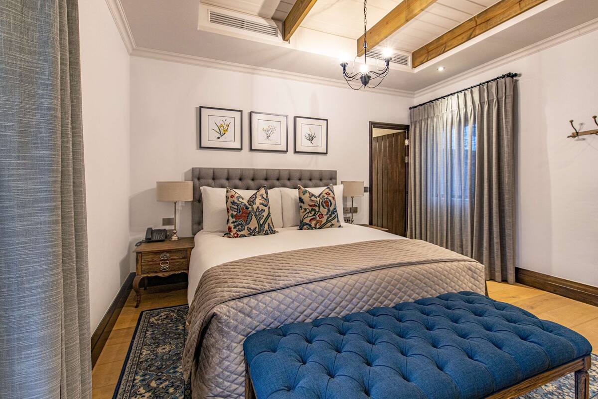 A bedroom with a large bed, a bedside table, a lamp, a blue ottoman, and a window with curtains.