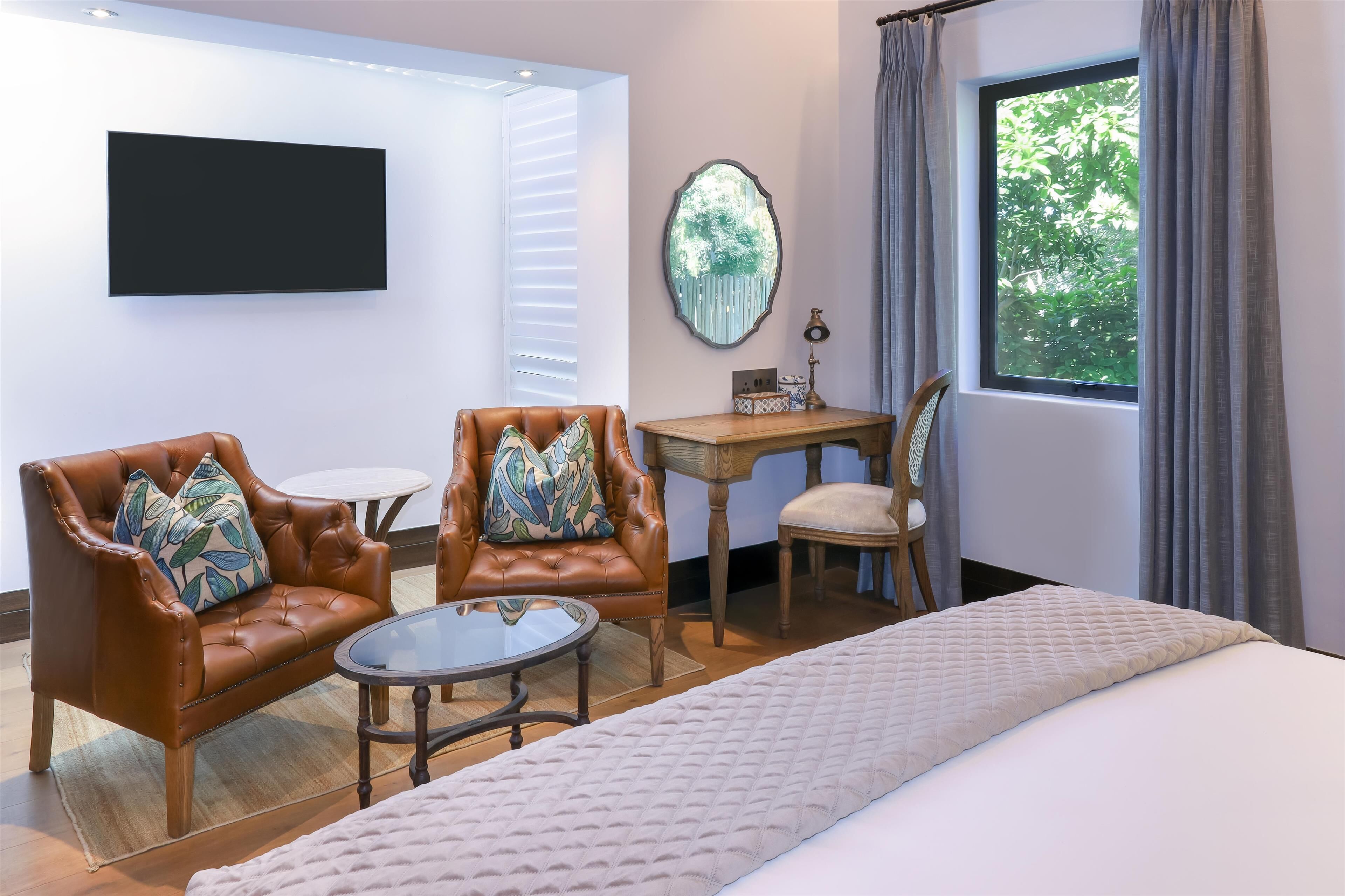 Een hotelkamer met een bed, twee leren fauteuils met gestreepte kussens, een klein rond tafeltje, een houten nachtkastje, een spiegel en een raam met gordijnen.