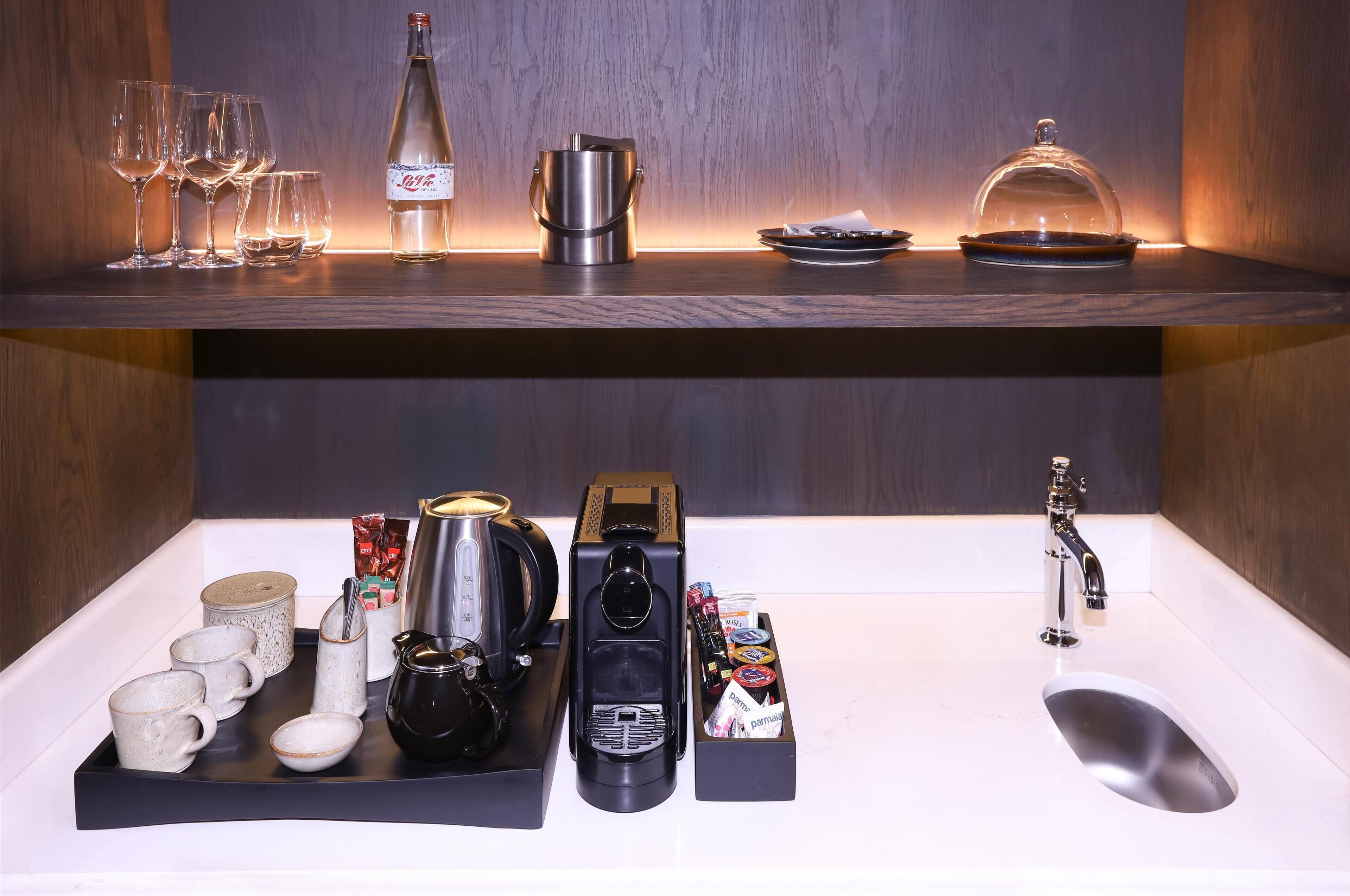 A hotel minibar with various items including glasses, a bottle, a coffee maker, and a tea set.