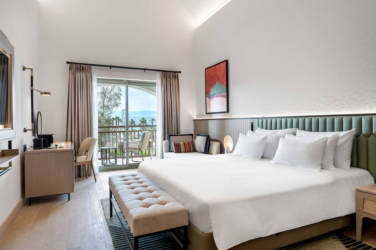 Una habitación de hotel moderna con una cama grande, un escritorio, un banco y un balcón con vistas.
