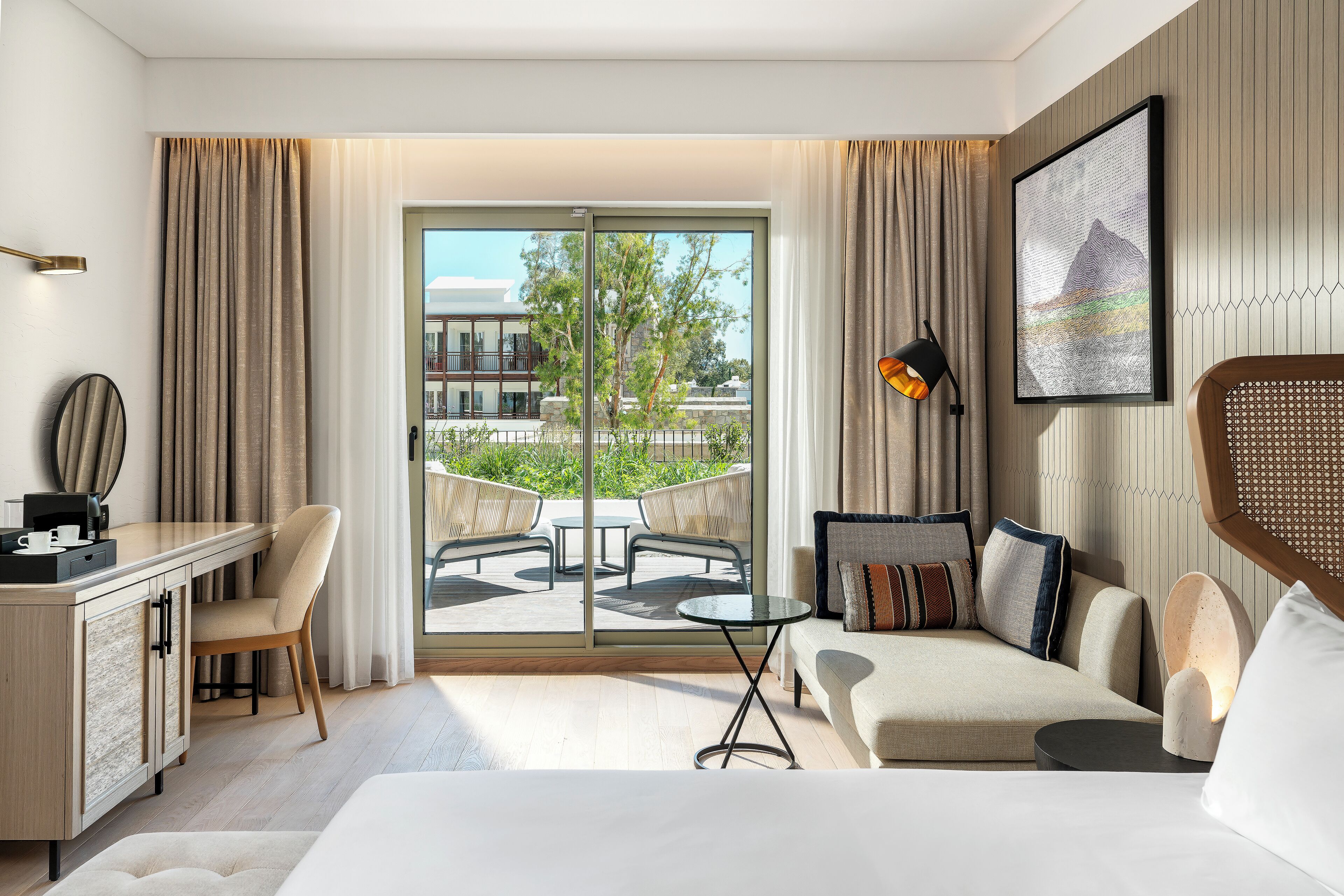 Une chambre d’hôtel moderne équipée d’un lit, d’un bureau, d’une chaise, d’un canapé, d’une petite table et d’un balcon avec mobilier d’extérieur.