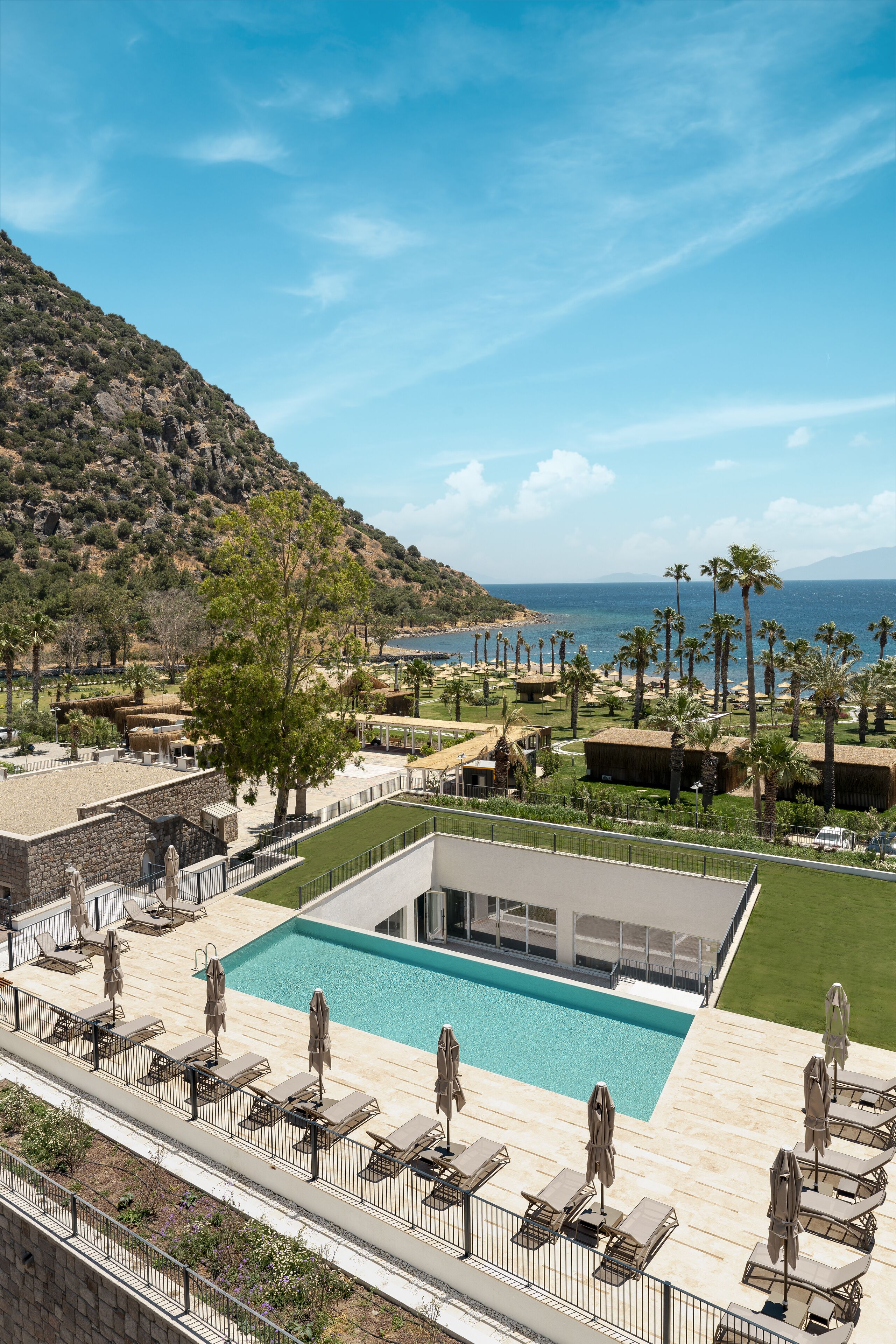 Una zona de piscina con sillones de descanso y sombrillas, un edificio con techo plano y vistas al mar y las colinas.