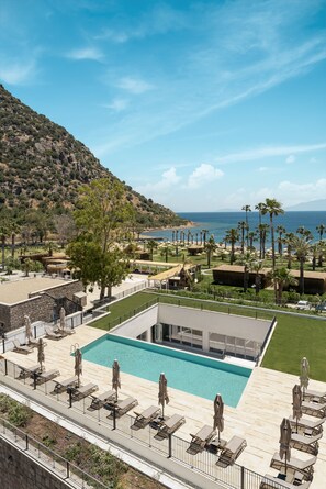A pool area with lounge chairs and umbrellas, a building with a flat roof, and a view of the sea and hills.
