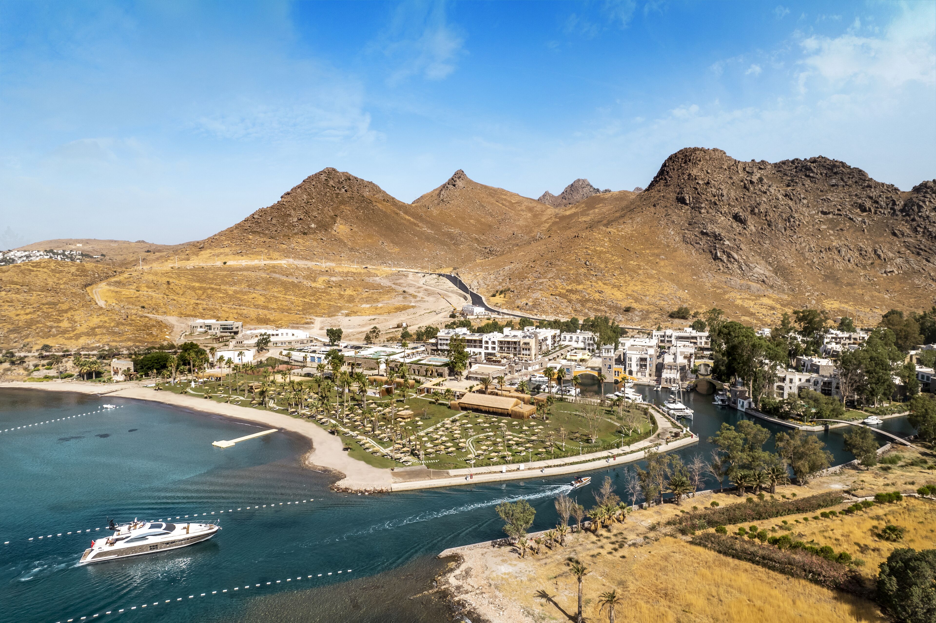 Un complejo turístico costero con un puerto deportivo, villas frente a la playa y montañas al fondo.
