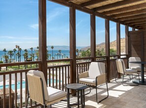 A covered outdoor seating area with a pool view, wooden beams, and a view of the sea and hills.