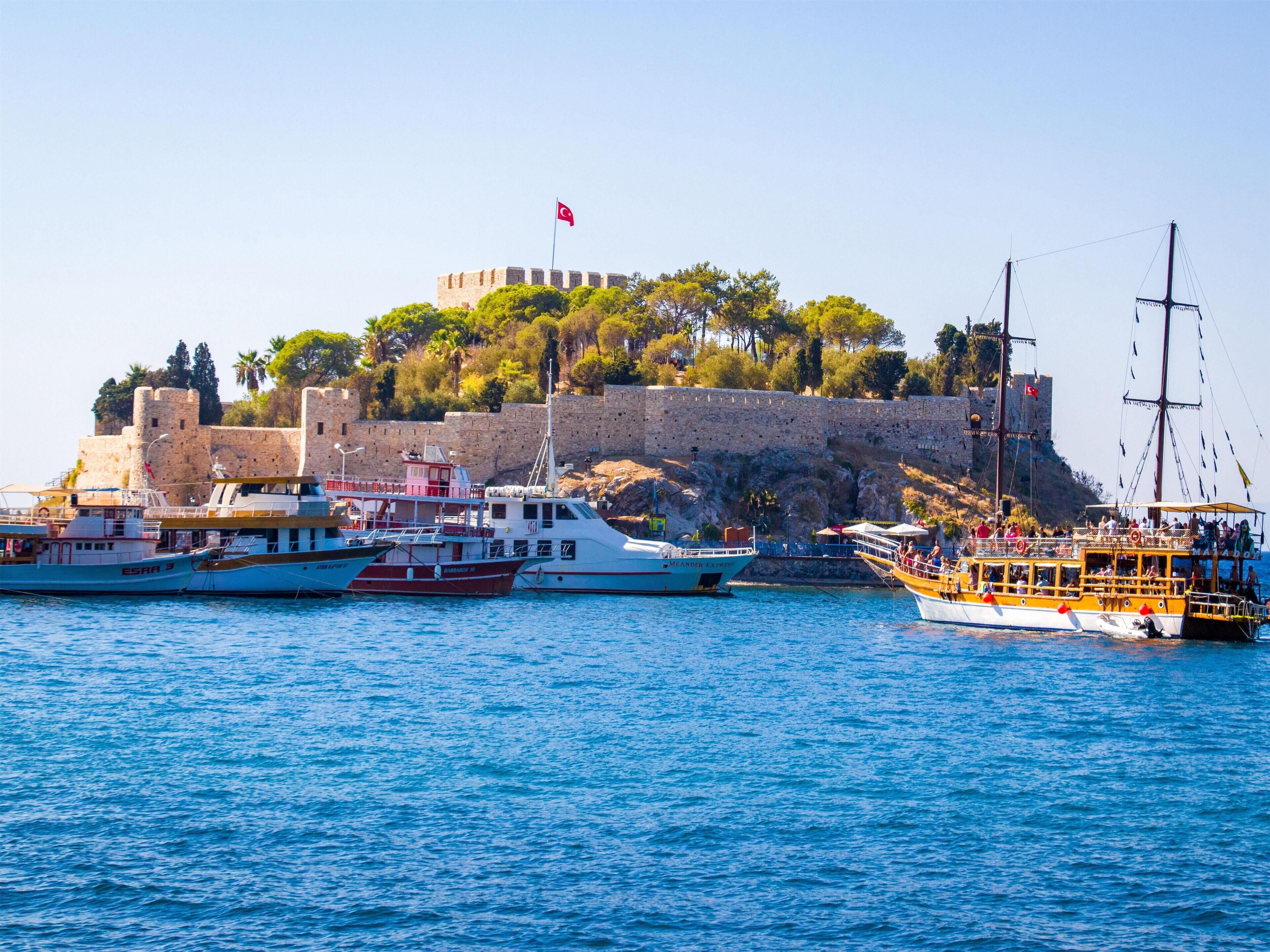A coastal scene with a historic castle, several boats docked in the harbor, and a clear blue sky.