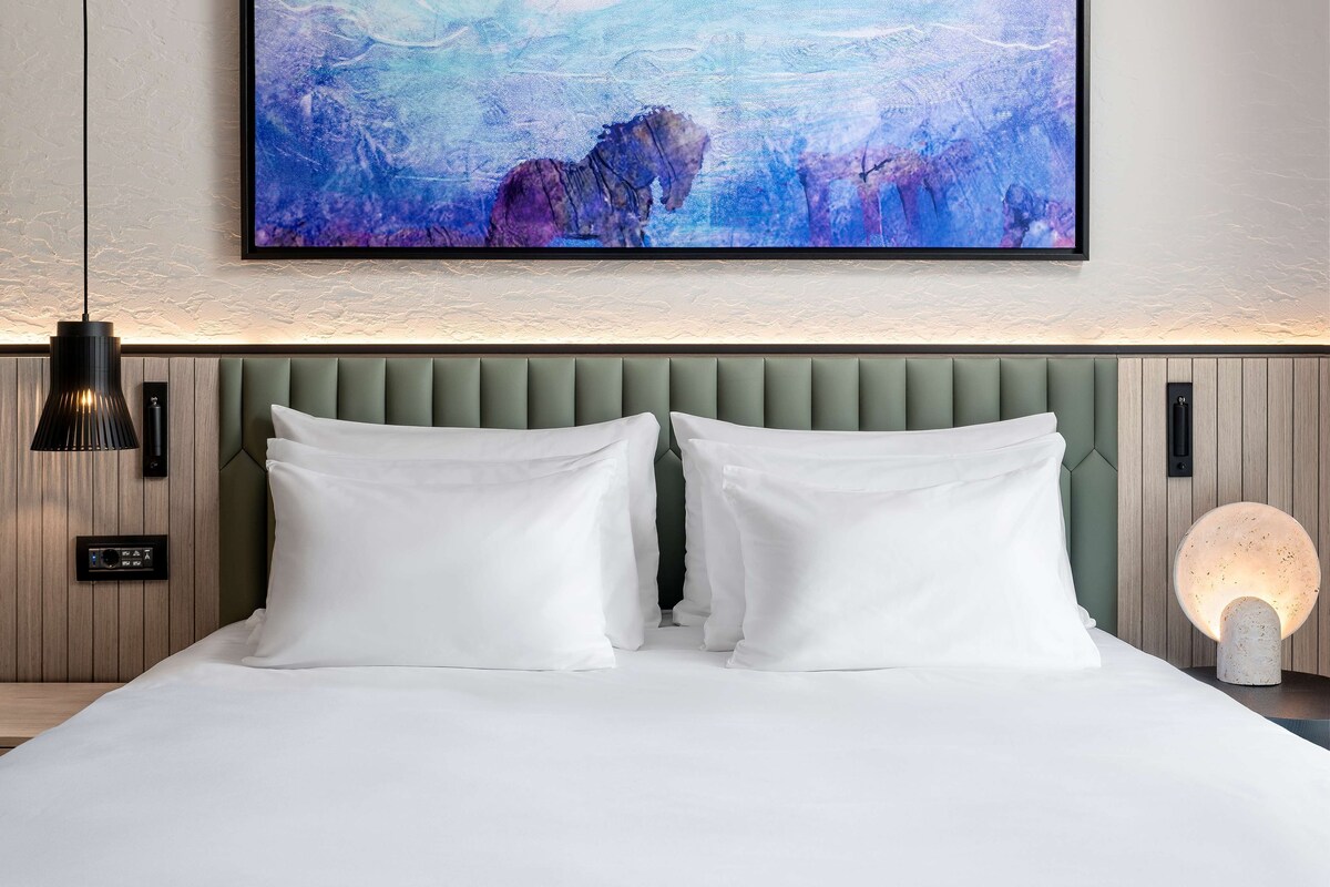 Une chambre d’hôtel avec un grand tableau abstrait, un lit moderne avec du linge de lit blanc, une tête de lit rembourrée de tissu vert et un luminaire fixé au mur.
