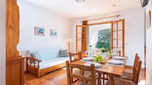 Un coin repas avec une table en bois, dressée pour quatre personnes, entourée de chaises. On aperçoit un canapé avec des coussins à l’arrière-plan.
