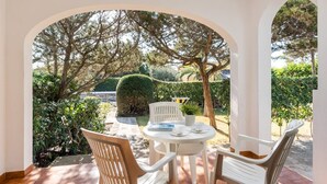 Une terrasse avec une table ronde et des chaises, entourée d’arches blanches, donnant sur un jardin avec des arbres et une haie soigneusement taillée.