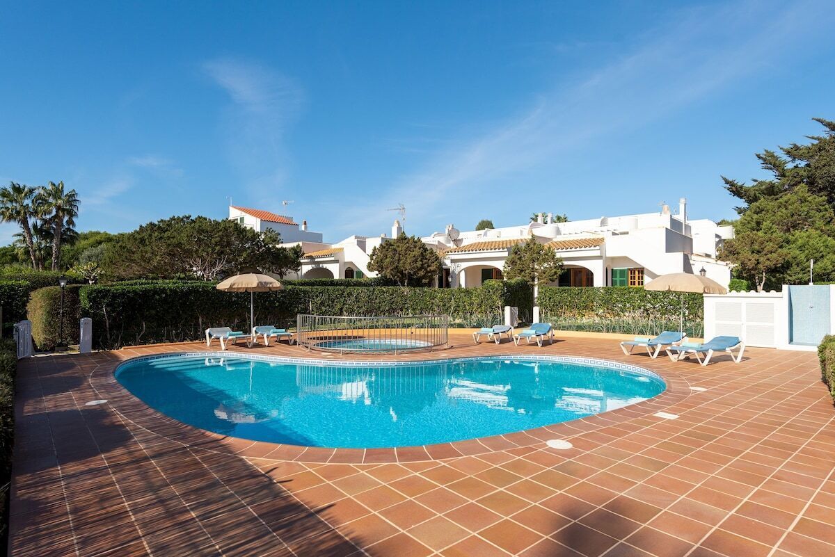 Ein Schwimmbecken mit Liegestühlen und einer gefliesten Terrasse, umgeben von Grünpflanzen und einem Gebäude mit weißen Wänden und roten Ziegeldächern.