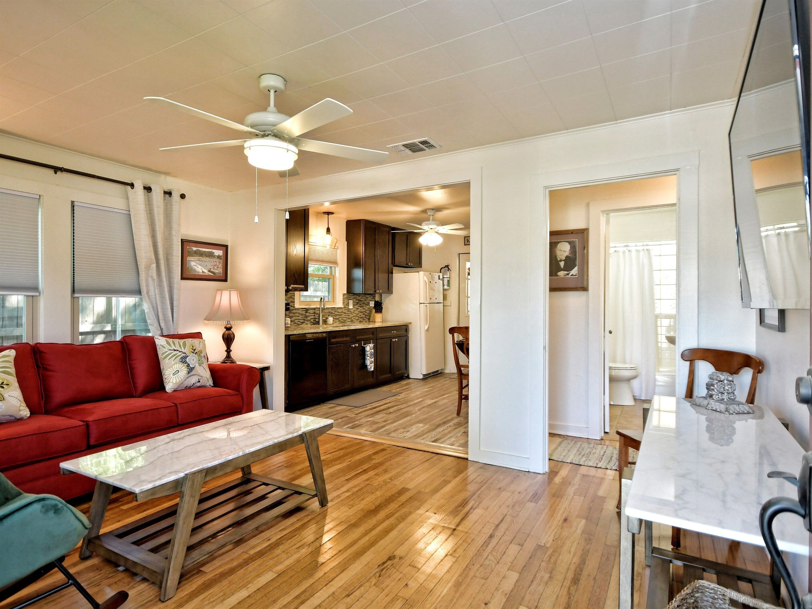 A spacious living area with a red sofa, a wooden coffee table, and a dining table with chairs. There is a kitchen in the background with a refrigerator and a microwave.
