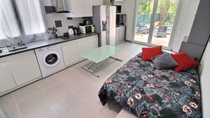 Un dormitorio pequeño con cama, una mesita y una lavadora en una cocina moderna con puerta corredera a un balcón.