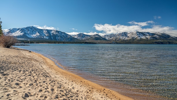 Ein Strand mit klarem Wasser, Bergen im Hintergrund und blauem Himmel.