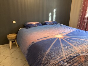 A bedroom with a bed featuring a sunset design, a small stool, and a plain wall.