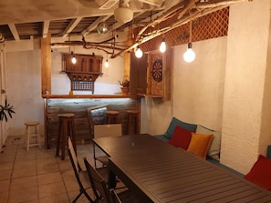A rustic interior with exposed wooden beams, a long wooden table with chairs, a dartboard, and a hanging light fixture.