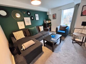 A living room with a dark sofa, a coffee table, and a blue armchair. There are wall decorations and a bookshelf. - Old Town Gem - Castle, Dark History, Uni (Edinburgh)