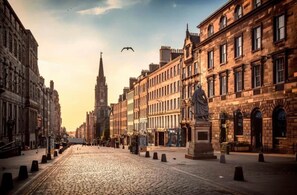 A cobblestone street lined with historic buildings, a prominent church spire, and a statue in the foreground.