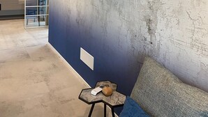 A modern room with a textured wall, a blue sofa, a small side table with a lamp, and a glass partition. - Maisonette (Tenno)