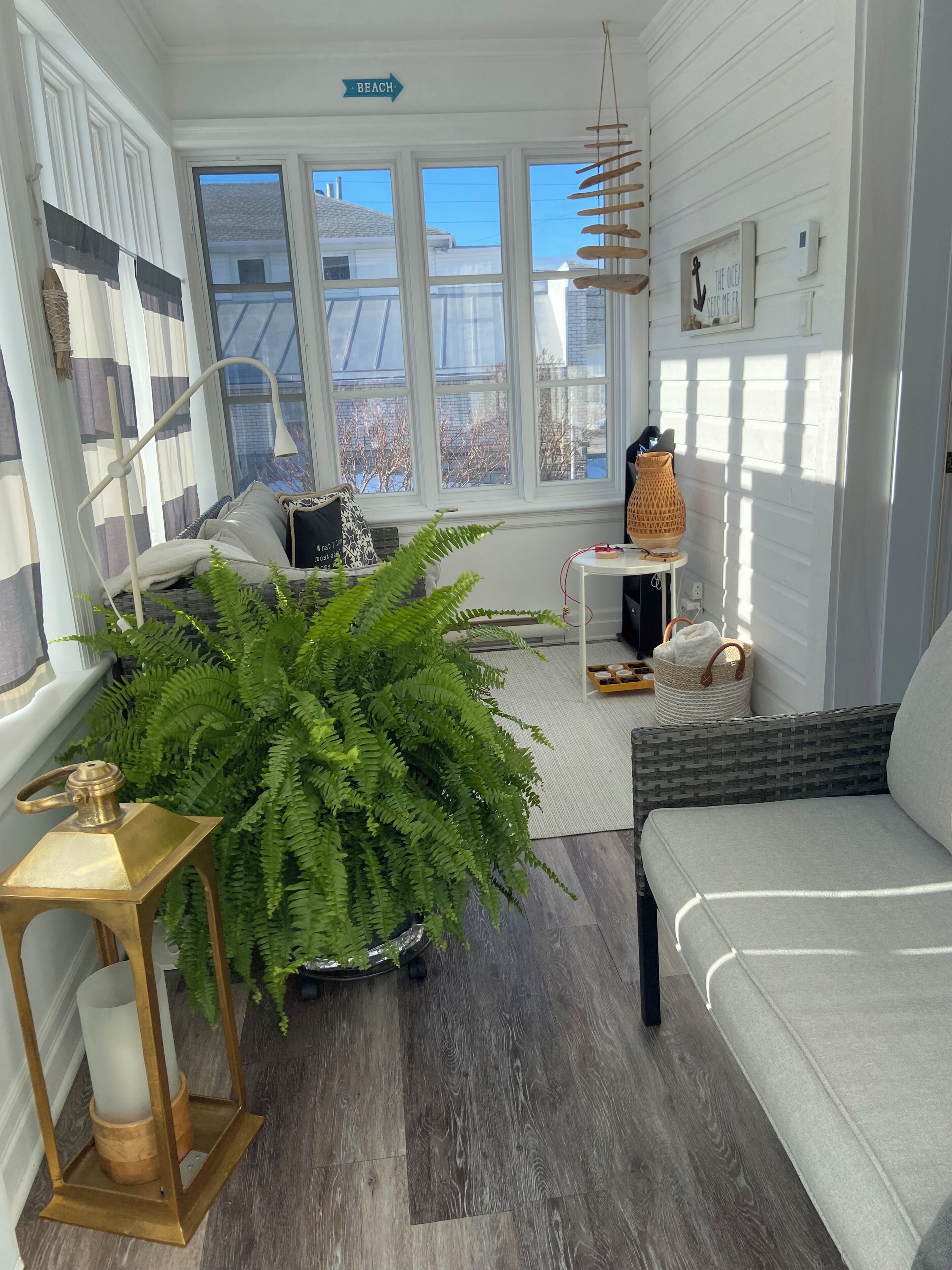 A cozy room with a large fern plant, a sofa, a lamp, and a sign indicating a beach direction.