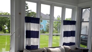 A cozy room with a white sofa, striped curtains, and a view of a blue house through large windows. - LA BELLE BLANCHE MAISON VUE DU FLEUVE, PRÈS DE MÉTIS SUR MER (Saint-Ulric)