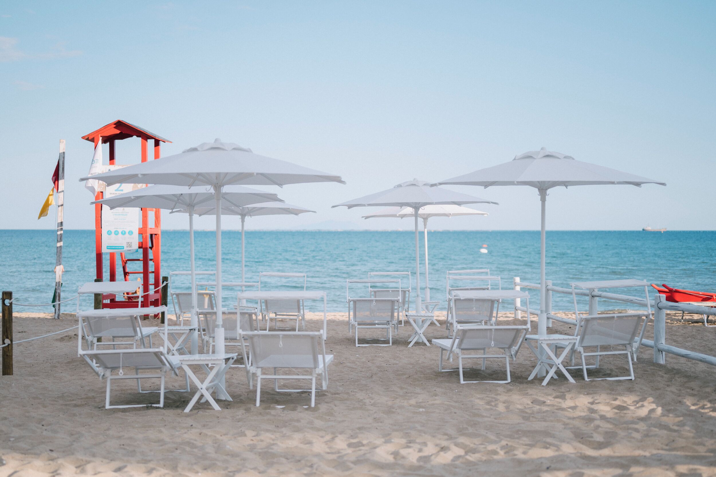 Une plage avec des chaises longues blanches et des parasols, une tour de surveillance pour les sauveteurs et un drapeau.