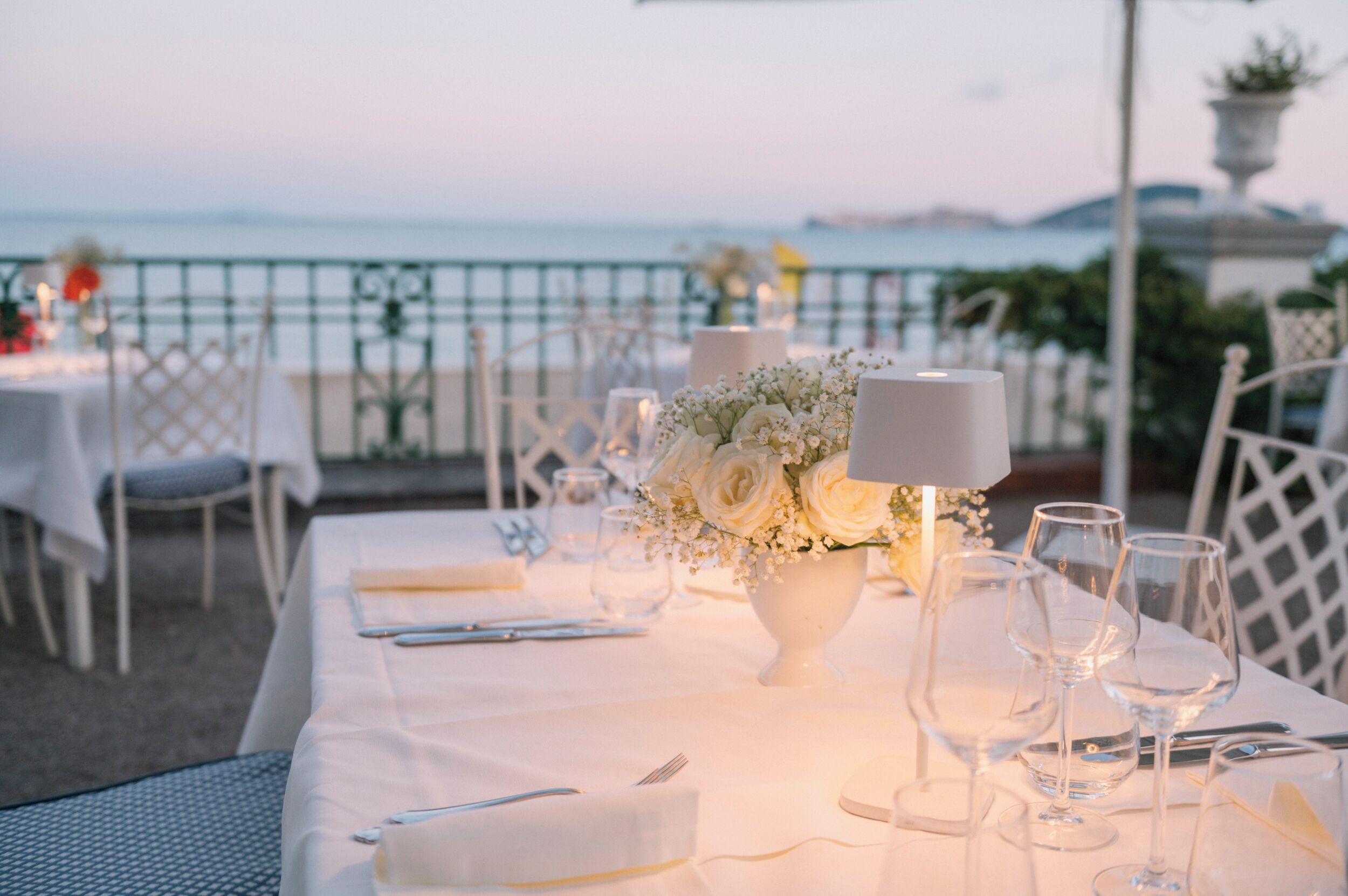 A table set for a meal with a view of the sea, white tablecloth, and a vase of flowers.