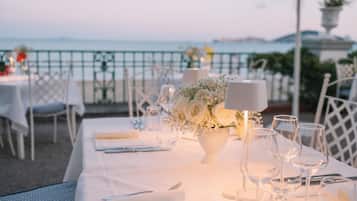 A table set for a meal with a view of the sea, white tablecloth, and a vase of flowers.
