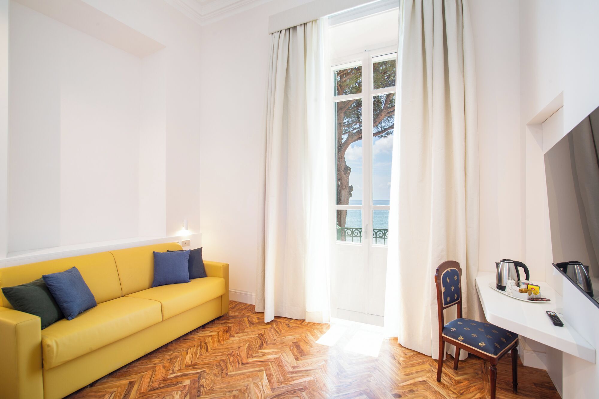 A bright room with a yellow sofa, a wooden herringbone floor, a white desk with a chair, and a view of the sea through a window.