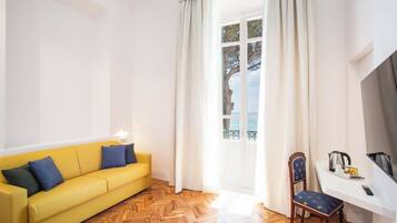 A bright room with a yellow sofa, a wooden herringbone floor, a white desk with a chair, and a view of the sea through a window.