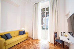 Um quarto claro com um sofá amarelo, piso de madeira em padrão herringbone, uma escrivaninha branca com cadeira e vista para o mar através de uma janela.