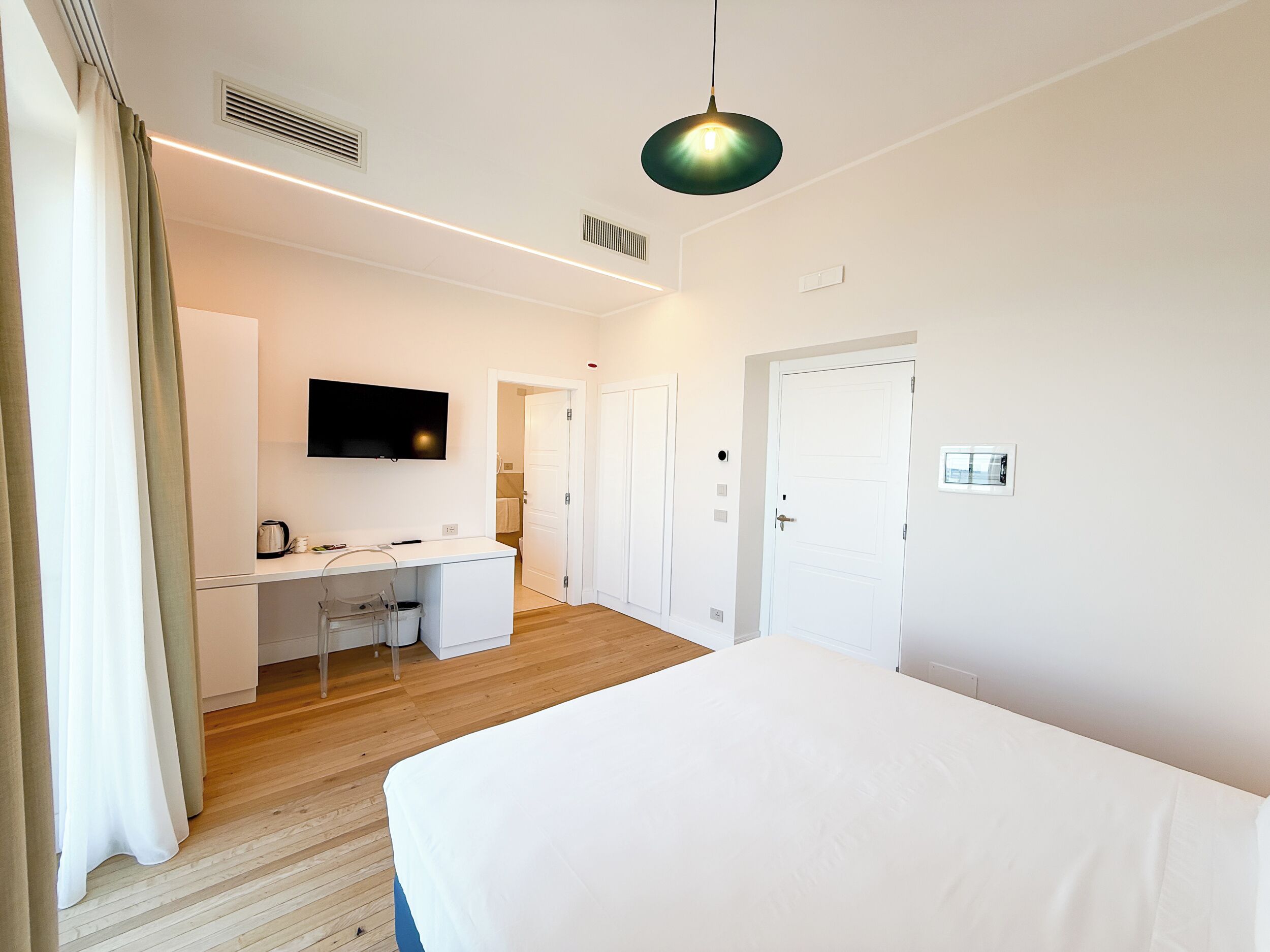 A modern hotel room with a bed, a desk with a chair, a television, and a door.