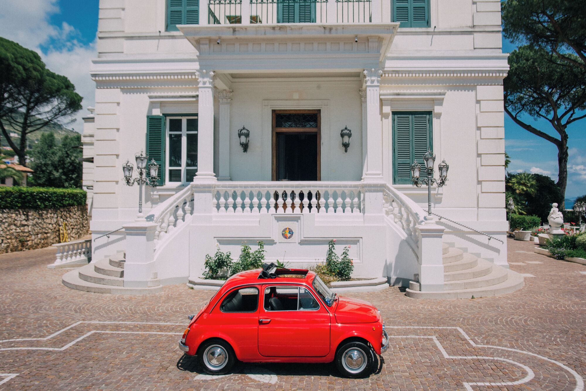 Un coche antiguo rojo estacionado frente a un edificio blanco con una entrada imponente y una escalera que lleva a un balcón.