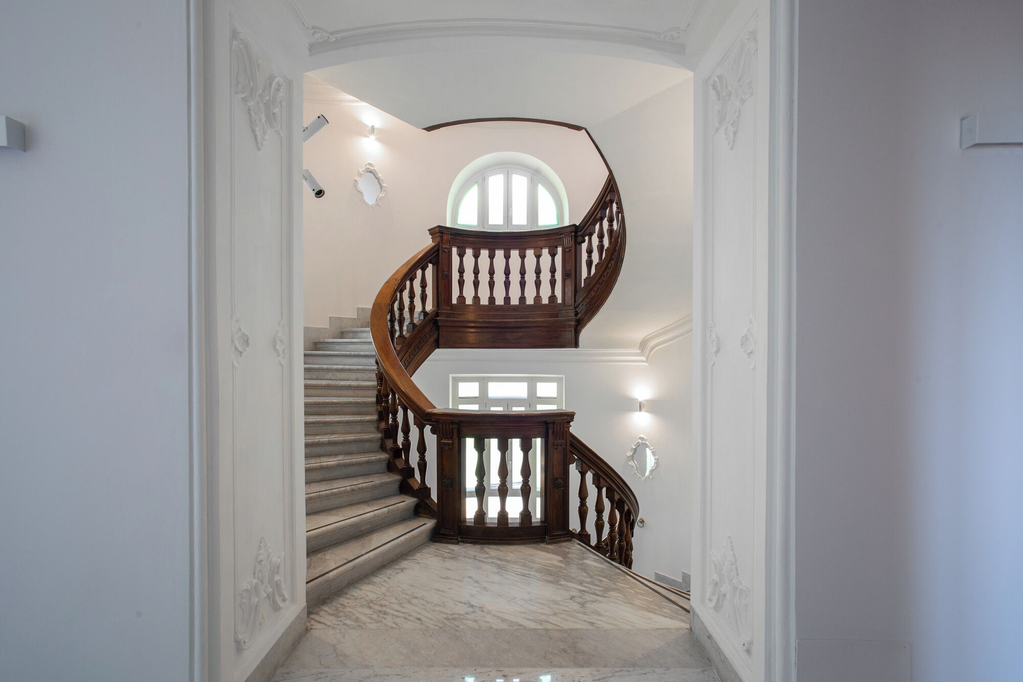 A grand staircase with a wooden handrail and marble steps, leading to a landing with a large arched window.