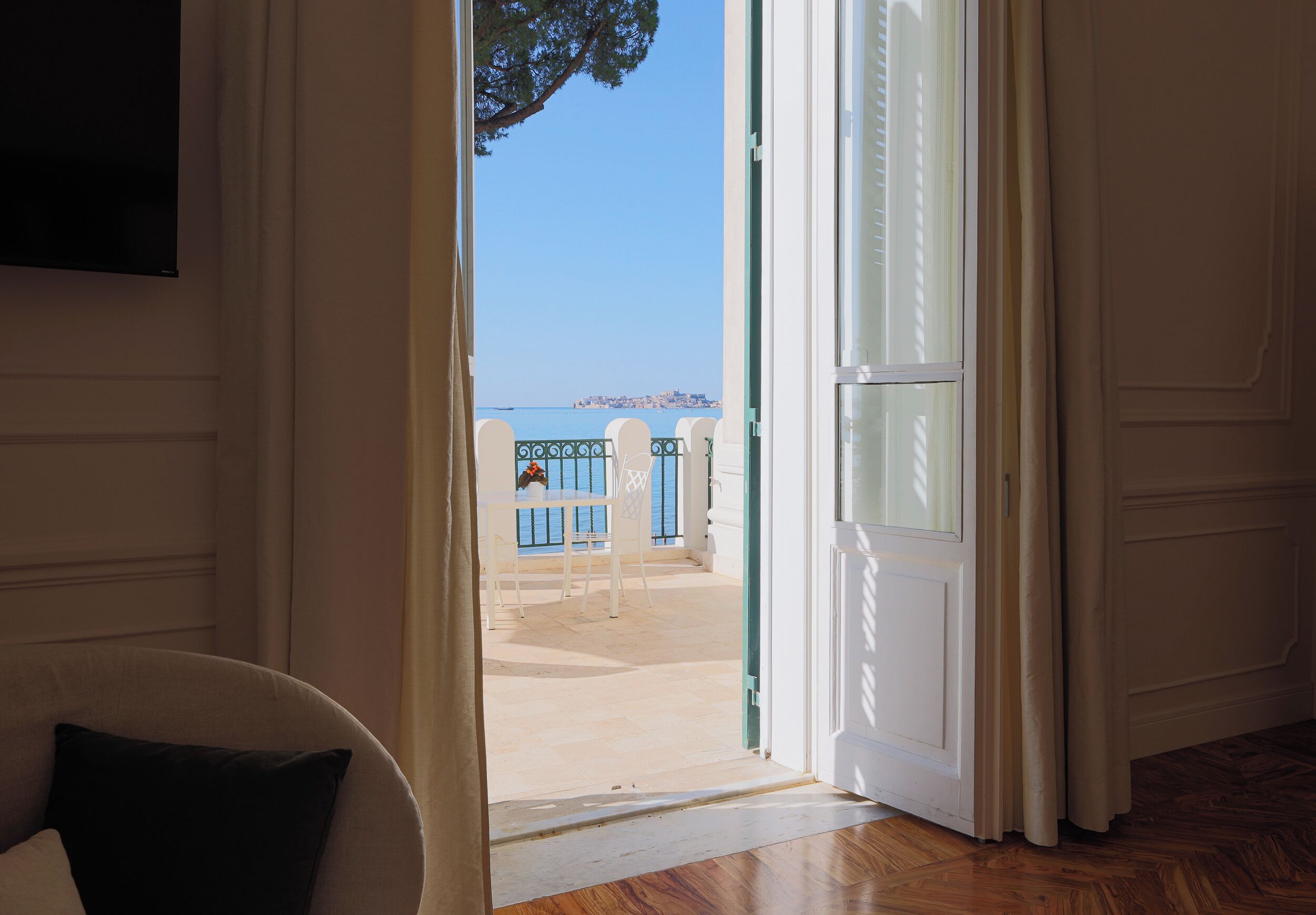 Eine geöffnete Tür, die auf einen Balkon mit Blick auf das Meer und eine entfernte Stadt führt.