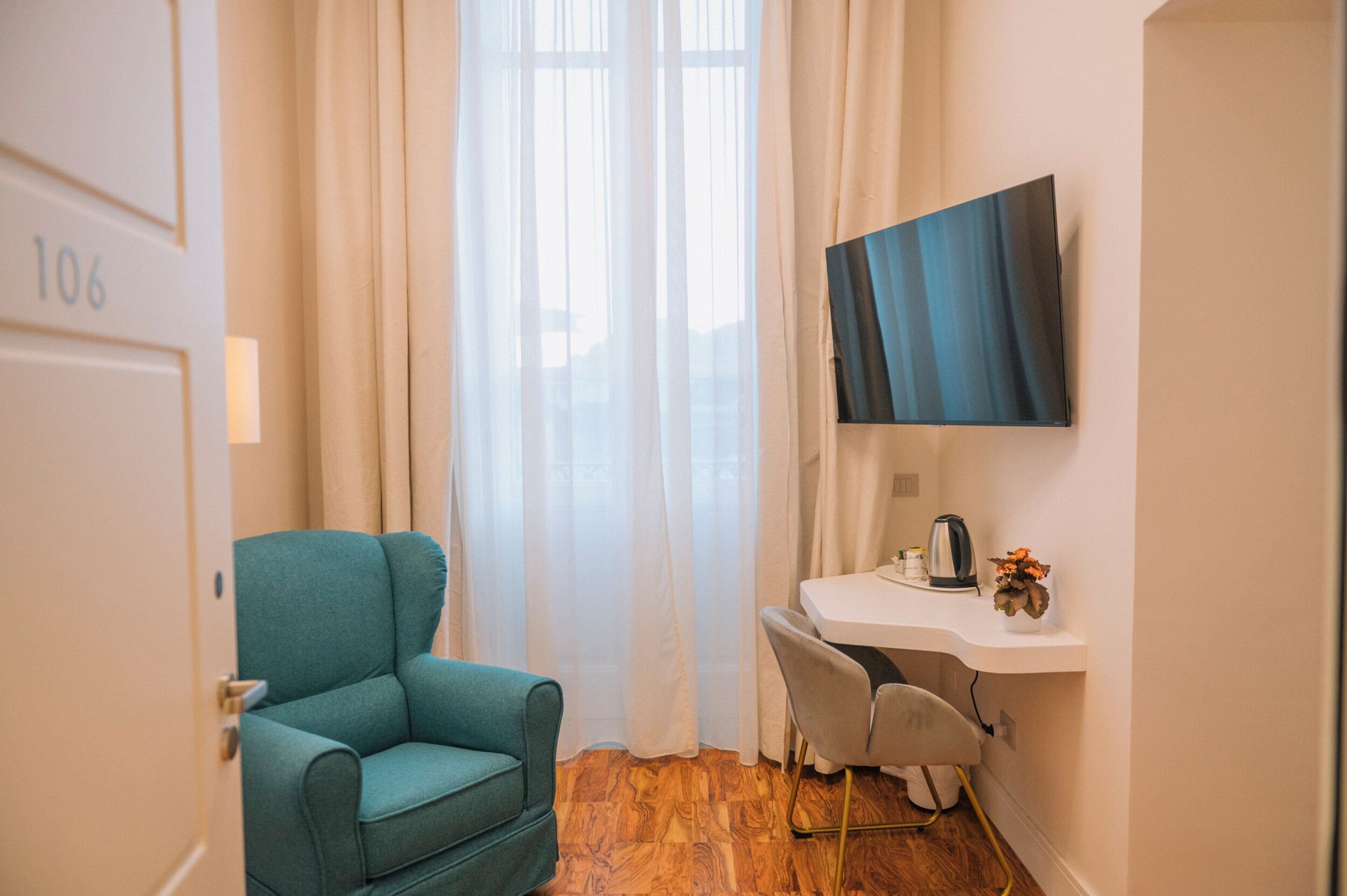 Una habitación con un sillón azul turquesa, un escritorio con televisor de pantalla plana y una ventana con cortinas traslúcidas.