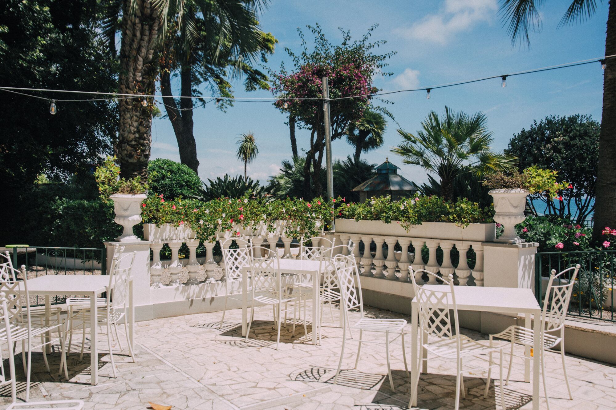 Un patio exterior con mobiliario de metal blanco, rodeado de macetas decorativas y diversas plantas tropicales.