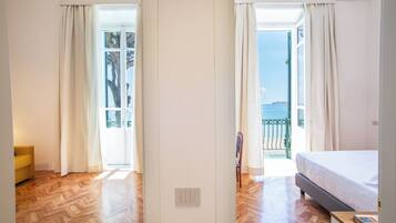 A bedroom with a bed, a wooden floor, and a view of the sea through an open window.