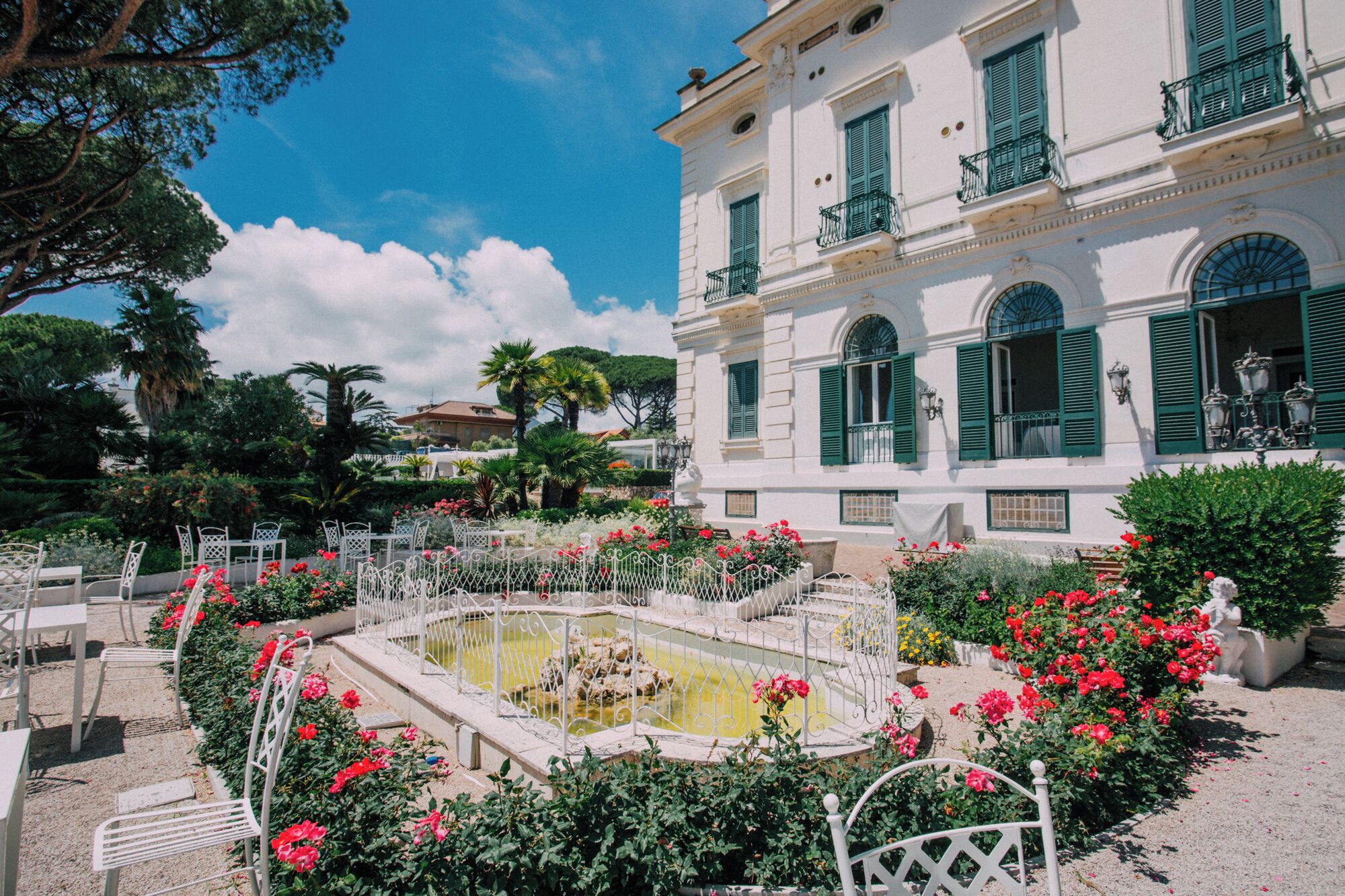 A courtyard with a fountain, surrounded by white metal chairs and tables, and a building with green shutters and balconies.